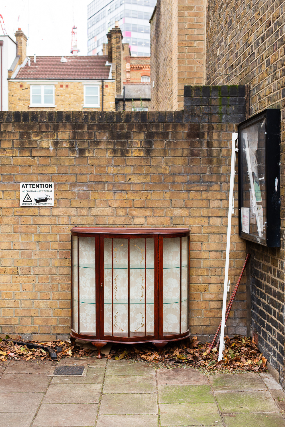Photograph of a vintage display cabinet made from wood and glass, with an ornate background interior, at the side of a street with a brick wall behind upon which is a sign that says 'ATTENTION,NO DUMPING or FLY TIPPING, OFFENDERS WILL BE PROSECUTED' with a sticker over the top saying 'DESTROY CAPITALISM BEFORE IT ESTROYS YOU'. To the right a noticeboard can be seen from the side with a white curtain rail leaning against it. Behind the wall there are houses and a tower block just visible and there is paved floor in the foreground with fallen leaves around the edges.