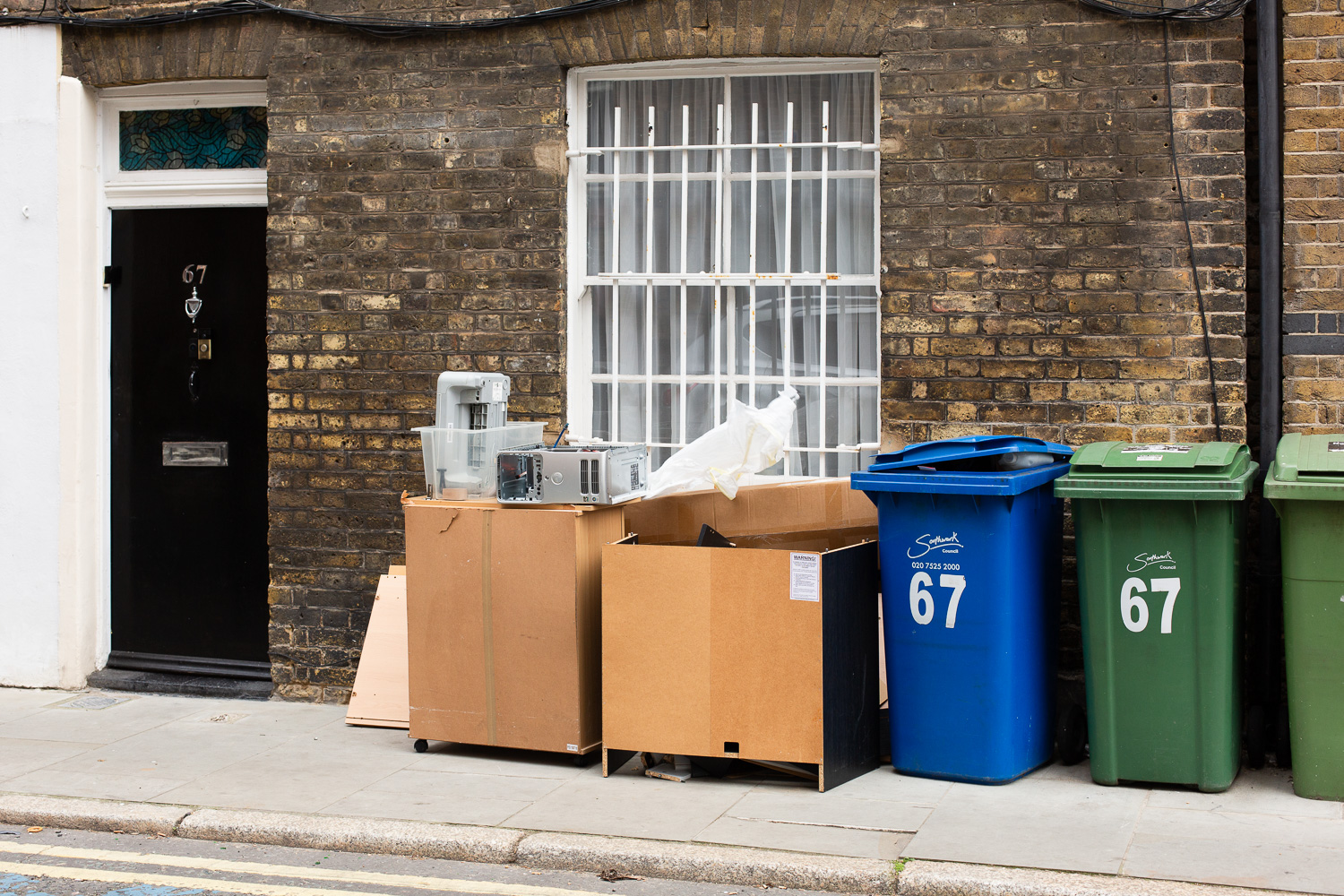 Photograph of a brick building with green and blue bins with '67' in large white numbers on the front, dismantled office furniture and equipment including a PC tower neatly lined up on the pavement next to them, in front of a window with white painted metal bars on the front. To the left is black door with a metal '67', door knocker and letter box,