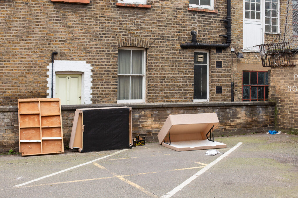 Photograph of the side of a carpark with broken bits of a dismantled bed with lift-up and drawer storage dumped against a low brick wall. Behind are windows and doors in a brick building and in the foreground are painted lines.