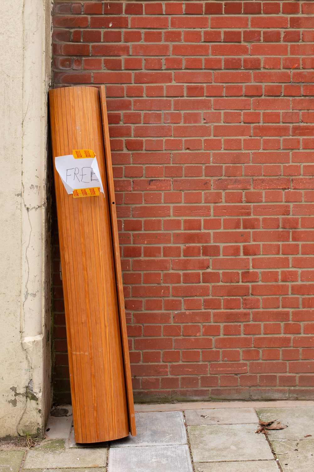 Photograph of some sort of folded up wooden screen with a piece of A4 paper taped to it with 'FREE' hand written in large letters. It is leaning against a cream wall, in front of a red brick wall and with pavind slabs below.