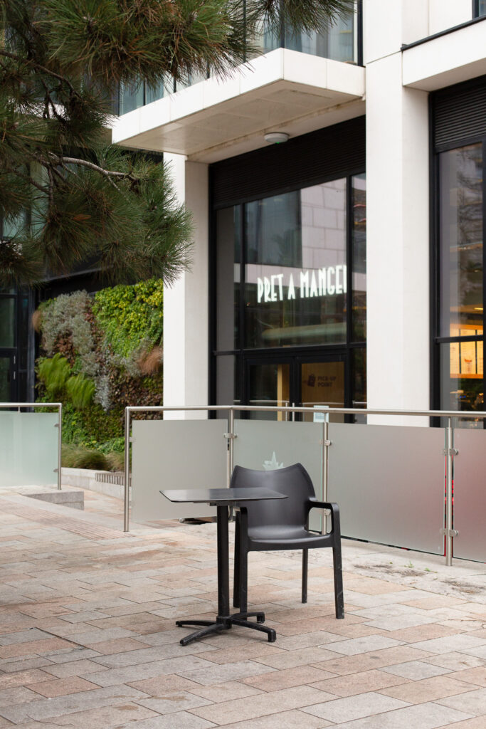 Photograph of a single black chair and small black table in a paved area with glass screening behind. In the background is a large glass contemporary building with 'PRET A MANGER' in illunminated white writing and a wall covered in plants behind. Part of a pine tree can be seen in the top left corner.