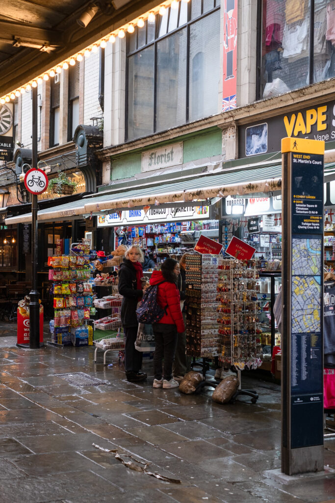 Photograph of a souvenir shop in london with two people browsing in front of displays of things such as fridge magnets. Above the canopy of the shop an old sign saying 'florist' is visible in old-fashioned writing. The paved floor is wet from rain, there's a sign indicating that bikes are not permitted to the left and to the right is a freestanding map and information board, above which can be seen the word 'VAPE' in yellow on the shop behind.