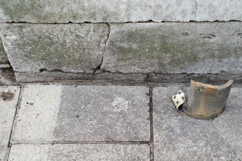 Photograph of an old wall and pavement with a piece of old pipe sticking up to the left with an old, worn 6 of spades card caught on it.