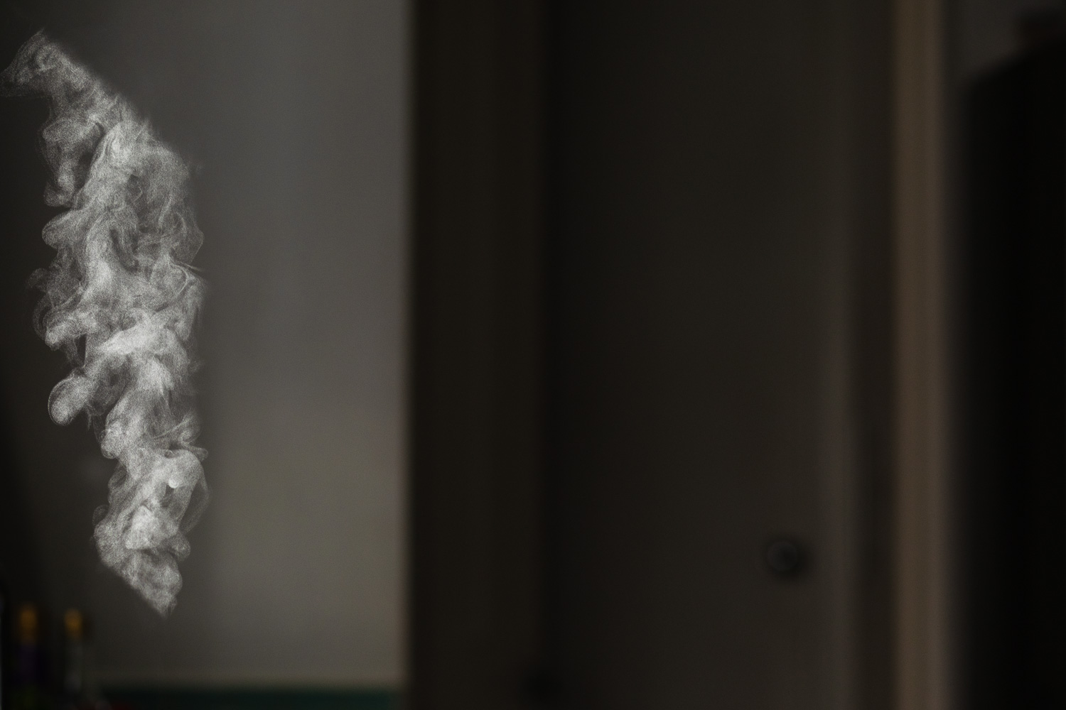 Photograph of a column of steam coming from a rice cooker that is out of frame below. The steam is caught in sunlight coming through a window to the left, highlighting the column and cut off at angles to the top and bottom. In the background is a dark room, you can just about make out the wall and a door set back to the right.
