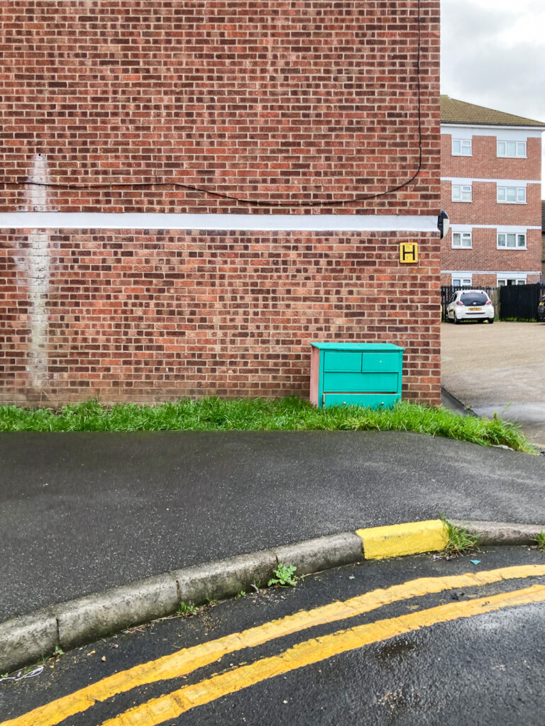 Photograph of a bright green painted chest of drawers on a piece of grass in front of the brick wall at the side of a block of flats, with a yellow fire hydrant 'H' sign and a strip of white above it, in front is a wide pavement and painted double yellow lines on a corner section of road and behind to the right is a carpark with a white car and more flats.