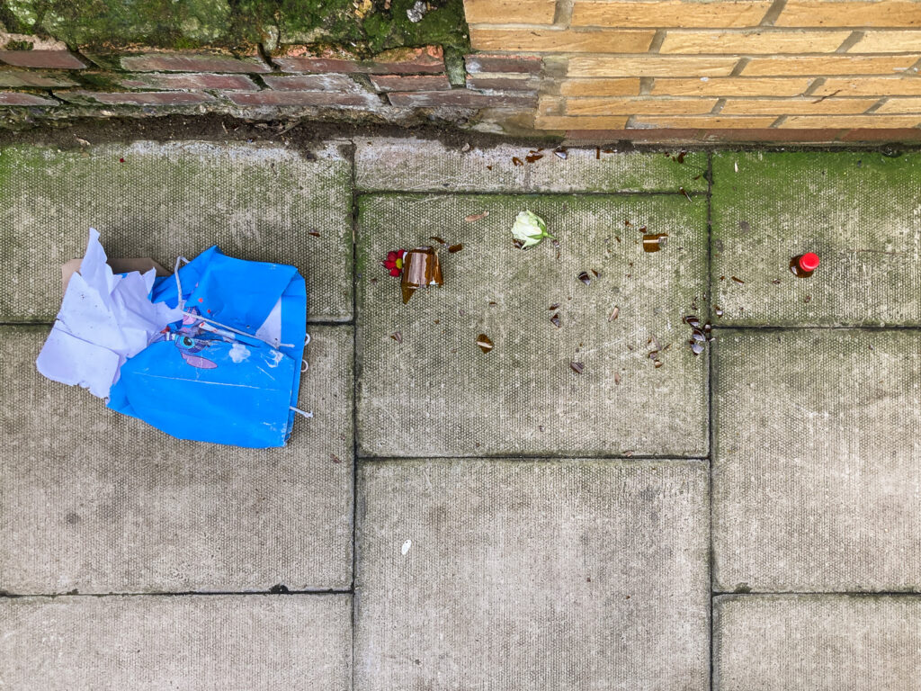 Photograph looking down at a paved pavement with a ripped blue Lilo and Stitch gift bag to the left, smashed brown glass bottle and a head of a white rose strewn across. Along the top of the frame a brick wall can be seen.