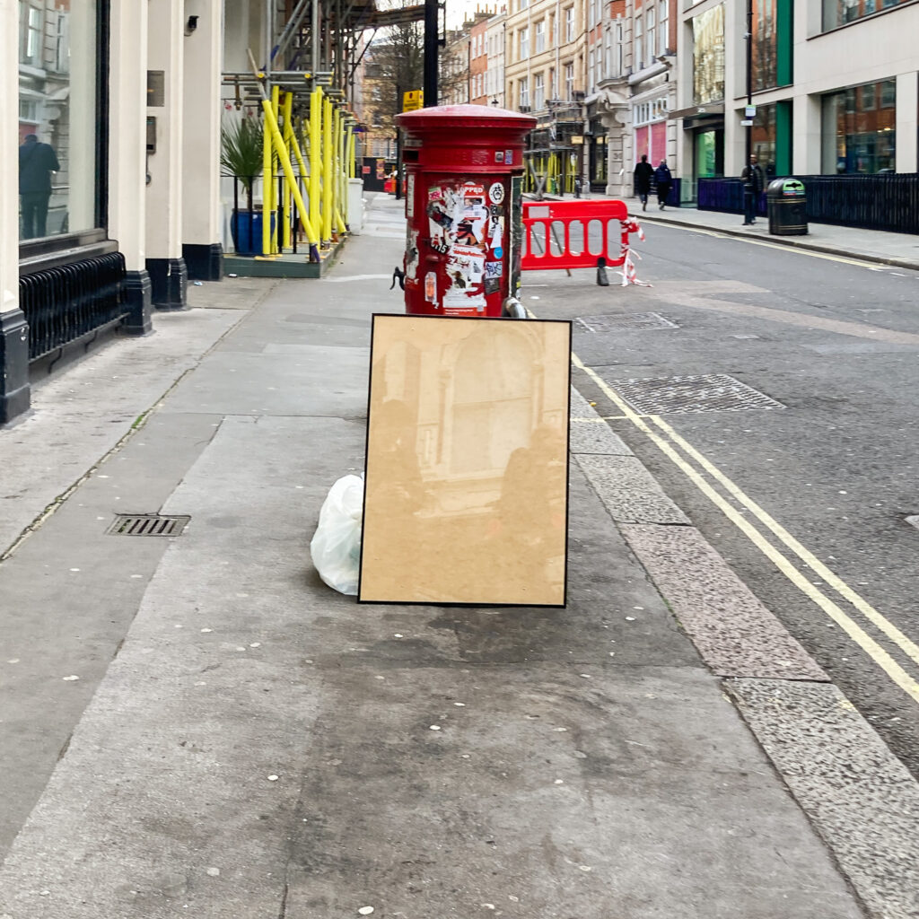 Photograph of an empty poster-sized frame leaning against bags of rubbish, in front of a red post box with stickers on, on a pavement with road to the right, shops and a red plastic barrier in the distance, and shops and scaffolding with yellow protective covering on it to the left.