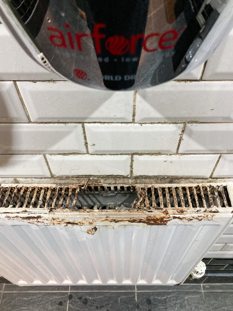 Photograph looking down at a white painted metal radiator with white metro tiles above and part of an 'airforce' hand dryer at the top. The radiator is rusty and has a hole in it, presumably where the water has been blown onto it.