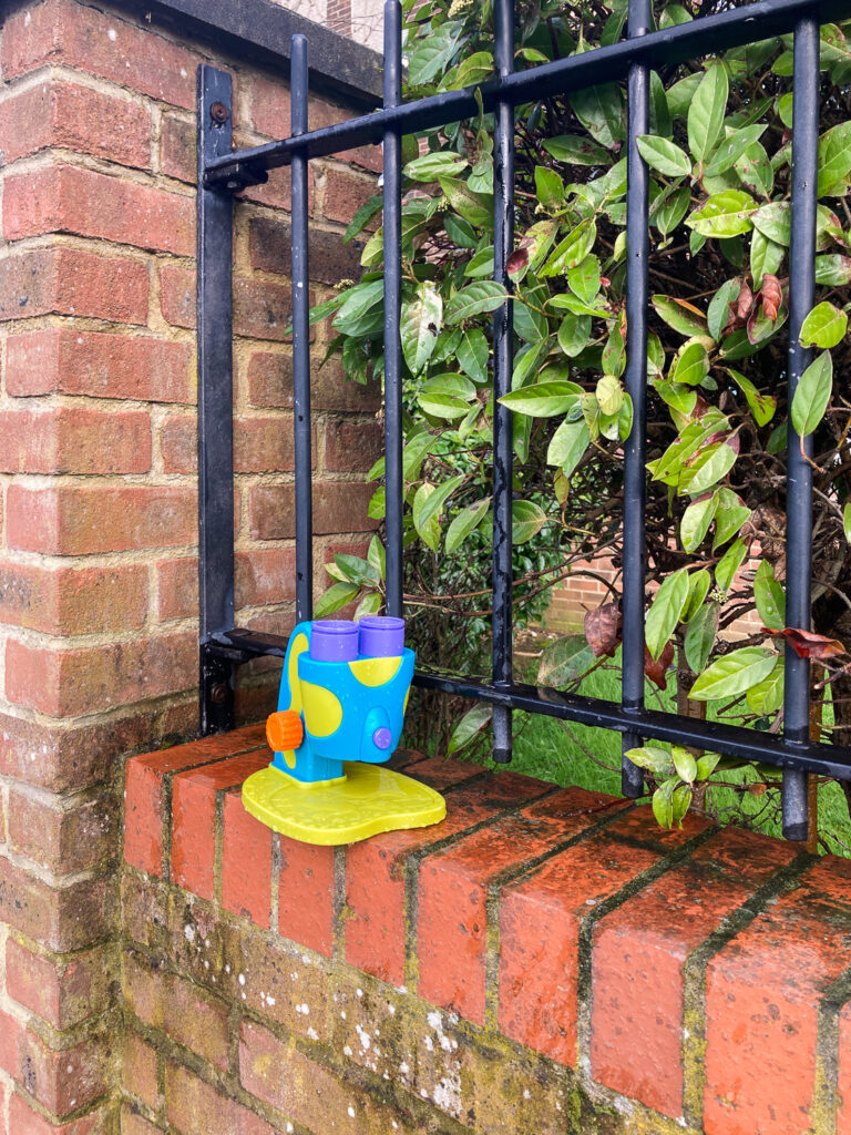 Photograph of a child's plastic toy microscope on a brick wall with black railings and a bush to the right.