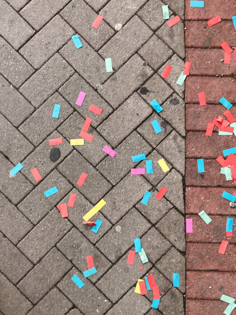 Photograph looking down at a pavement made from bricks in a pattern arrangement, with colourful pieces of large rectangular confetti scattered across it.