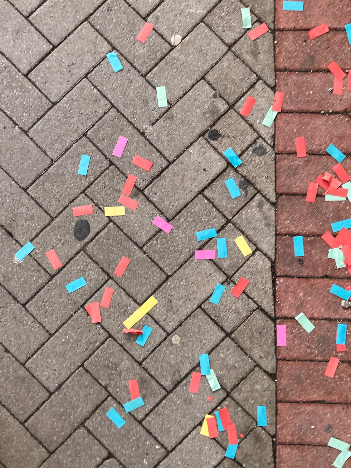 Photograph looking down at a pavement made from bricks in a pattern arrangement, with colourful pieces of large rectangular confetti scattered across it.