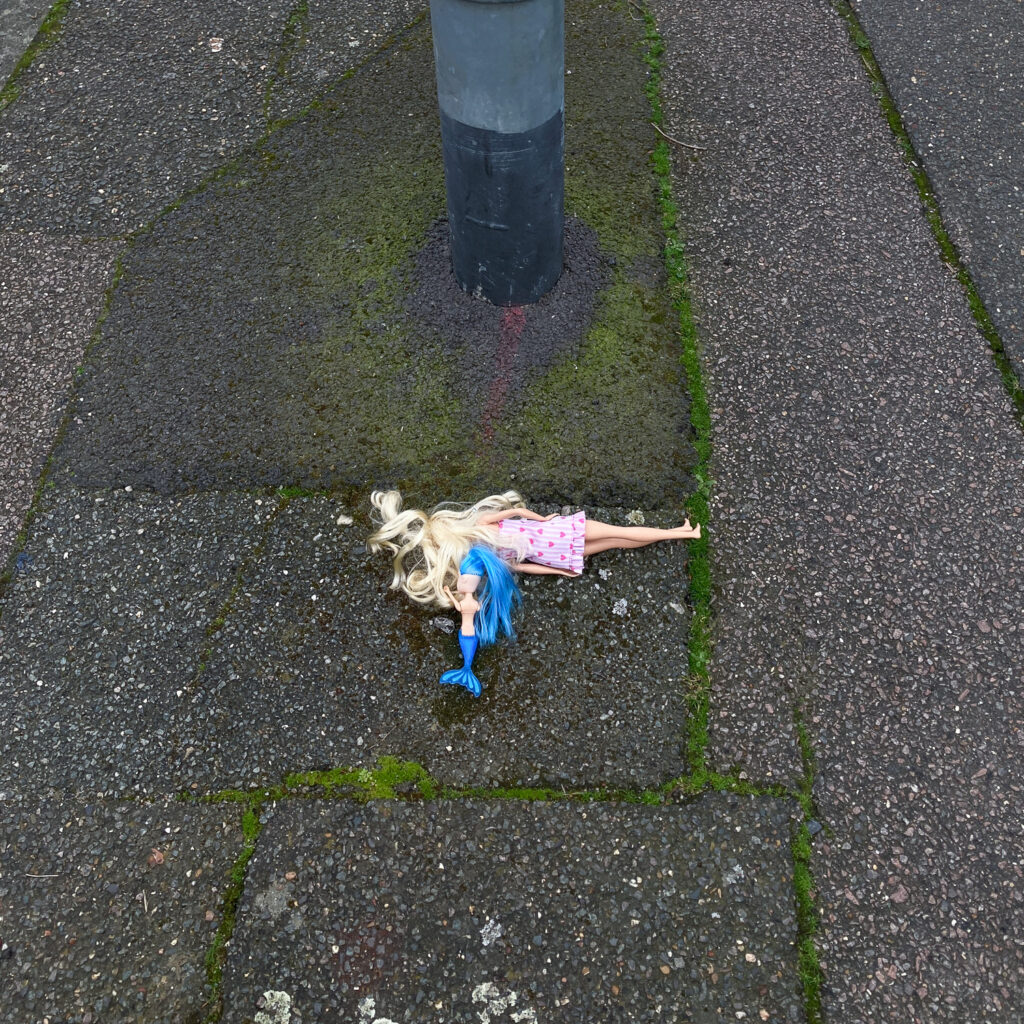 Photograph of a blonde Barbie-style doll in a pink dress with a blue-haired and blue-tailed mermaid doll. Both are face down on a pavement, which has moss growing in long lines of cracks and the bottom of a metal pole visible at the top of the frame.