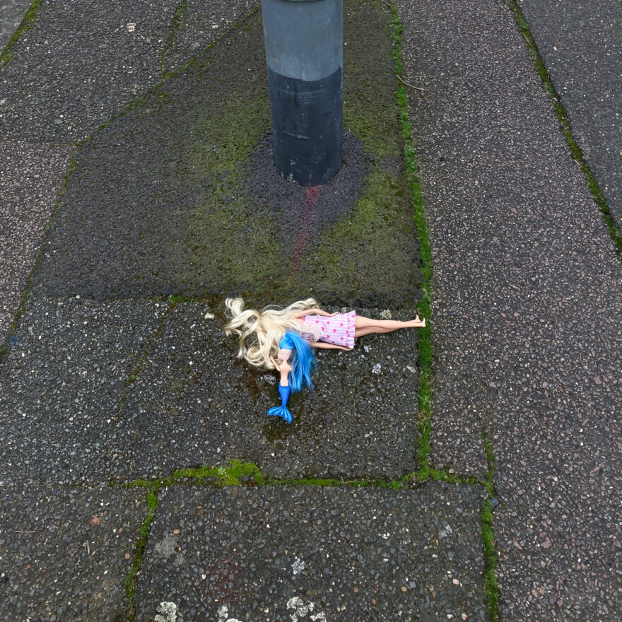 Photograph of a blonde Barbie-style doll in a pink dress with a blue-haired and blue-tailed mermaid doll. Both are face down on a pavement, which has moss growing in long lines of cracks and the bottom of a metal pole visible at the top of the frame.