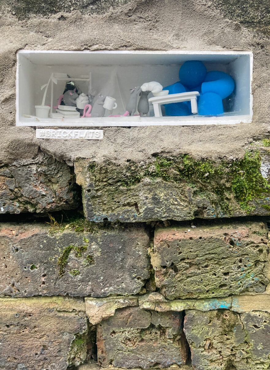 Photograph of a brick wall with a white recessed area cemented in at the top, with a scene of small mice and a large blue character having a tea party, and BRICKFLATS written underneath.