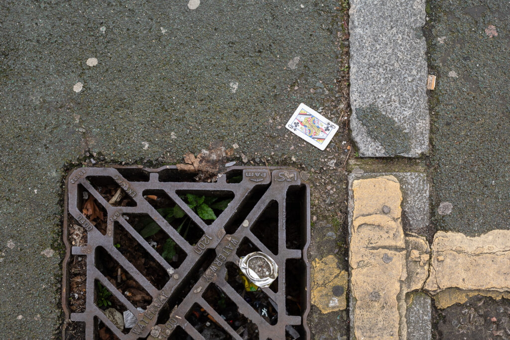 Photograph looking down at a Queen of clubs playing card on the floor, with a drain at the bottom of the frame, and paving and paint elements to the right.