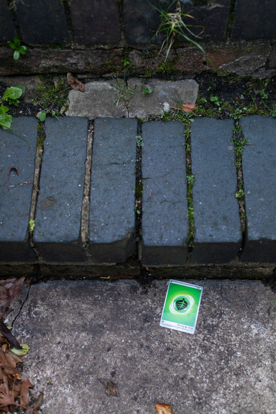 Photograph of a Pokémon Card on the floor in front of a brick step. The card is upside down, green and has 'Basic Engergy, ENERGY, Basic Energy' written on it and a graphic of a leaf in the centre.