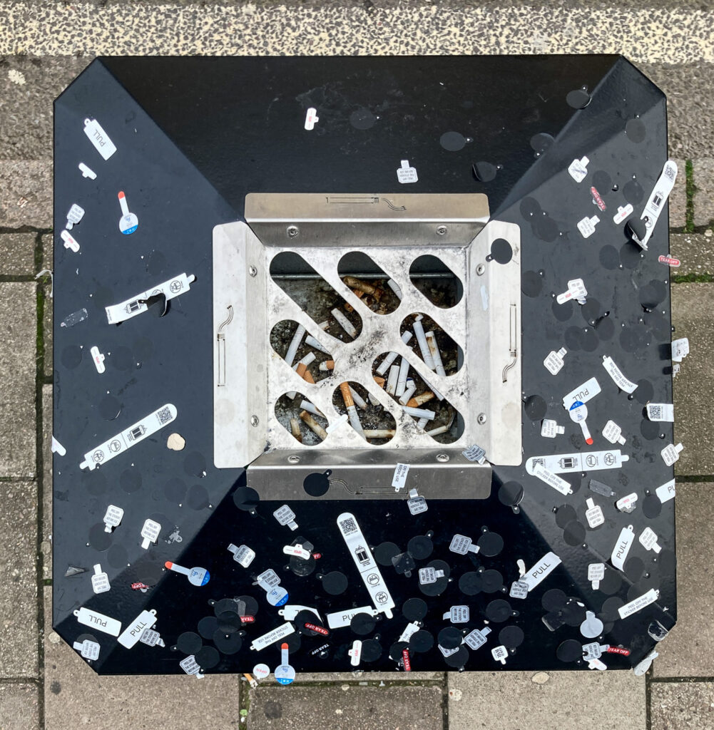 Photograph looking down on the top of a bin in a street, with an ashtray filled with cigarettes in the centre, and the black surround is covered in stickers from vape packets. The paved floor is visible around the edges behind.