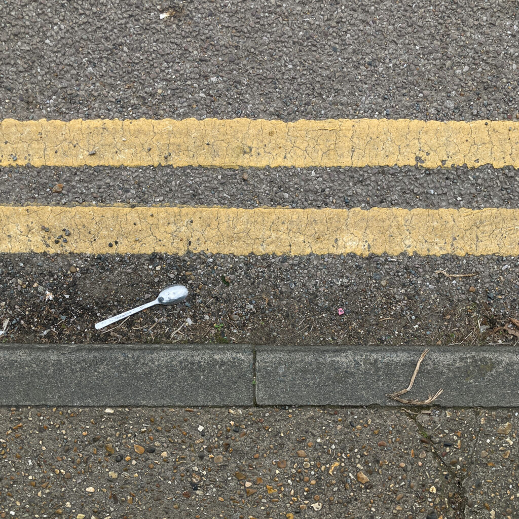 Photograph looking down at the curb at the side of a road, with pavement at the lower part of the frame, a metal spoon face down to the left above that, and double yellow painted lines above.