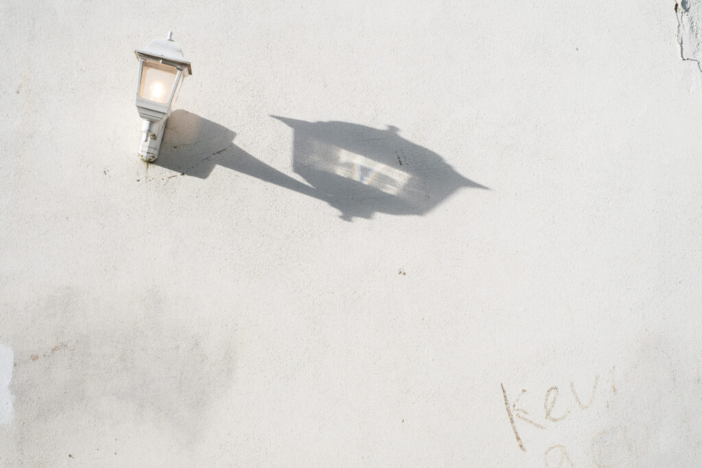 Photograph of a painted white wall with an old-fashioned white lantern on the wall to the left, with a warm lightbulb on. The lantern is tilted to the right and a long shadow from it extends to the right. At the bottom right are handwritten letters partially rubbed out, 'Kevin... a cu...