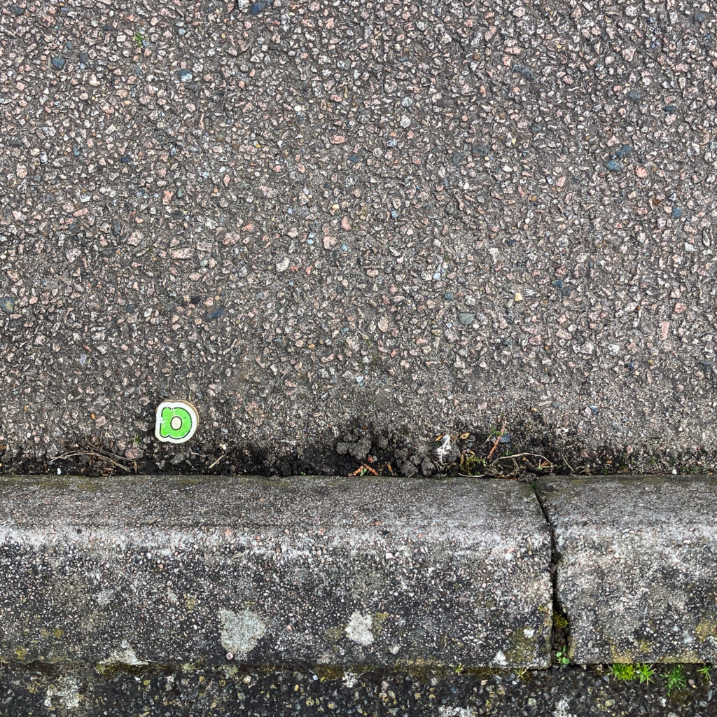 Photograph looking down at the curb at the side of a road, with pavement at the lower part of the frame, road above that and a wooden child's toy with a lower case letter 'a' in green on it, and road filling the top portion of the frame.