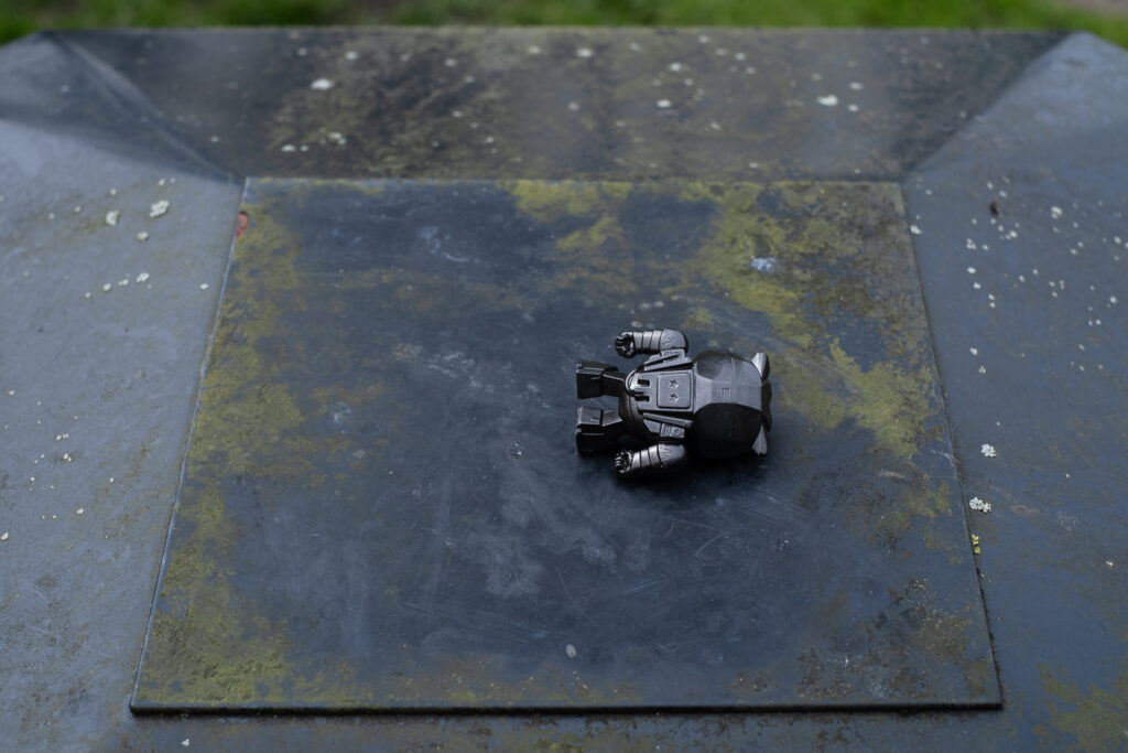 Photograph of a child's black robot toy face down on the top of a black metal bin, which has green and grey lichen on it.