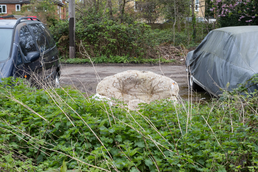 Photograph of the top of an old-fashioned upholstered armchair visible behind a hedge of nettles and long dried branches, between two parked cars (the one to the right covered), with more greenery and a wooden post visible in the background.