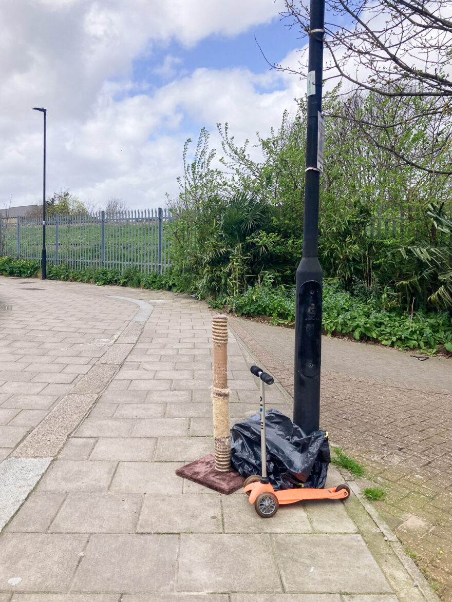 Photograph of a paved area with a cat scratch pole, a black bin bag and a child's scooter by a black lamppost. In the distance is an area of plants, a metal security fence and another black lamppost.