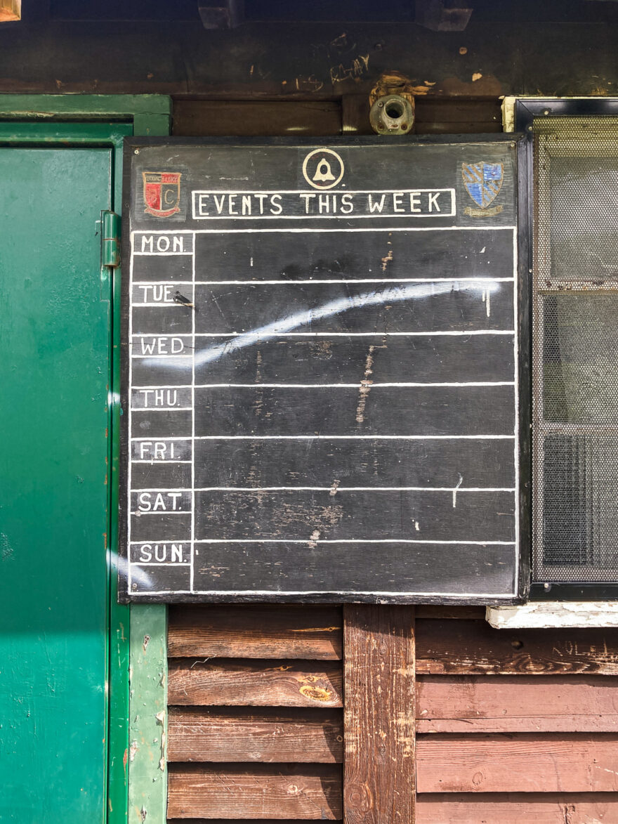 Photograph of a black board with 'EVENTS THIS WEEK' and some logos on it, with the days of the week 'MON, TUE, WED, THU, FRI, SAT, SUN'  and empty spaces to the right for things to be written in. The board is mounted on a wooden building with a covered window to the right and a green door to the left.