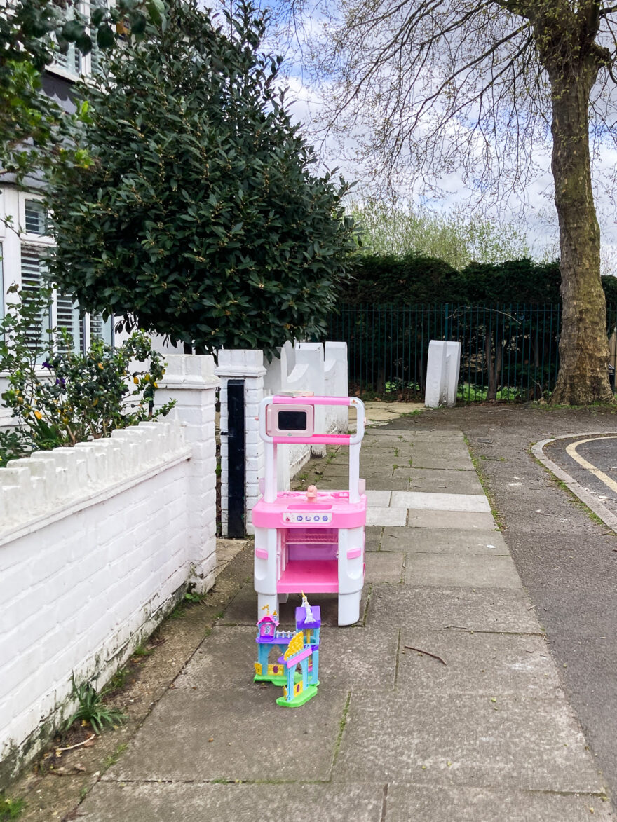 Photograph of a paved area with a plastic pink child's cooker and a small castle toy outside a house with white painted brick low wall, a tree, hedge and fence in the background and edge of the road to the right.