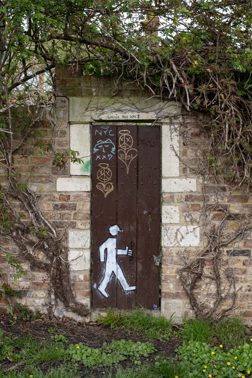 Photograph of a brick wall with a brown gate in the centre, with grafitti of a person walking and opening a handle at the bottom, hearts, a cat and 'NYc' toward the top, and 'NARNIA THIS WAY' with a downward pointing arrow in black on the wall above the gate. Below is grass and weeds and above are branches and climbing plants.