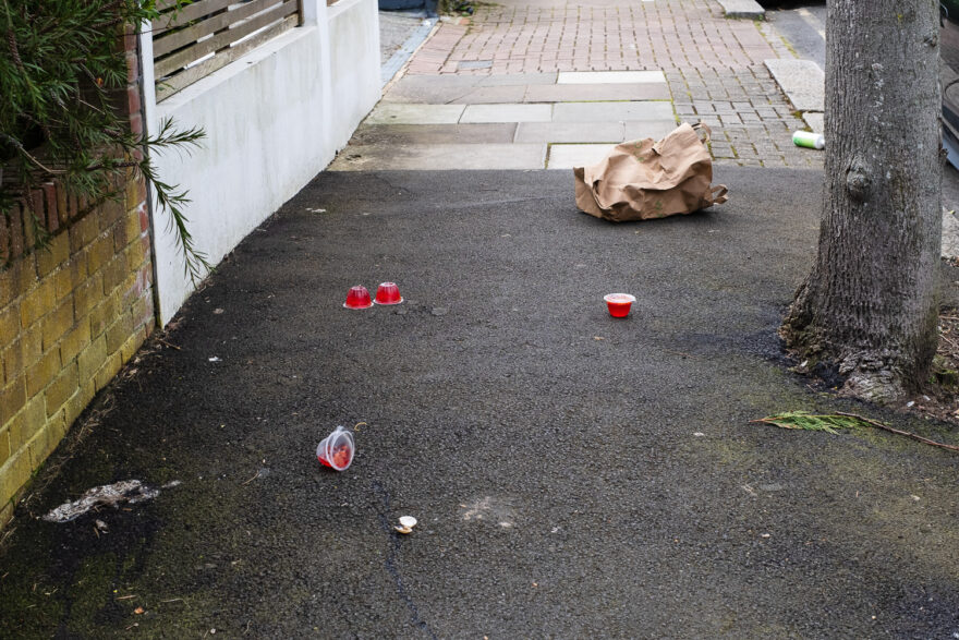 Photograph of a pavement with a large tarmac section upon which are scattered 4 pots of red jelly and a paper bag behind. To the right is a tree trunk and to the left are walls lining the path - one brick and one white. In the background the continuing path is paved with large slabs and then smaller bricks.