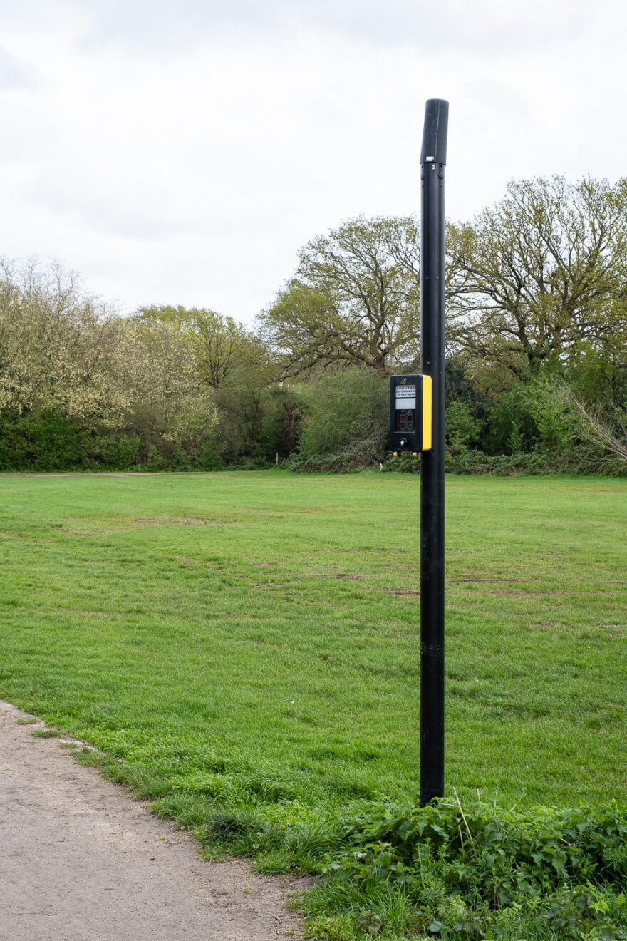 Photograph of a black pole with an equestrian crossing box halfway up, to be pressed by people riding horses who wish to cross the road. Behind is a grassy field with trees and bushes in the distance and a cloudy sky above and in the bottom left corner is a portion of dusty path.