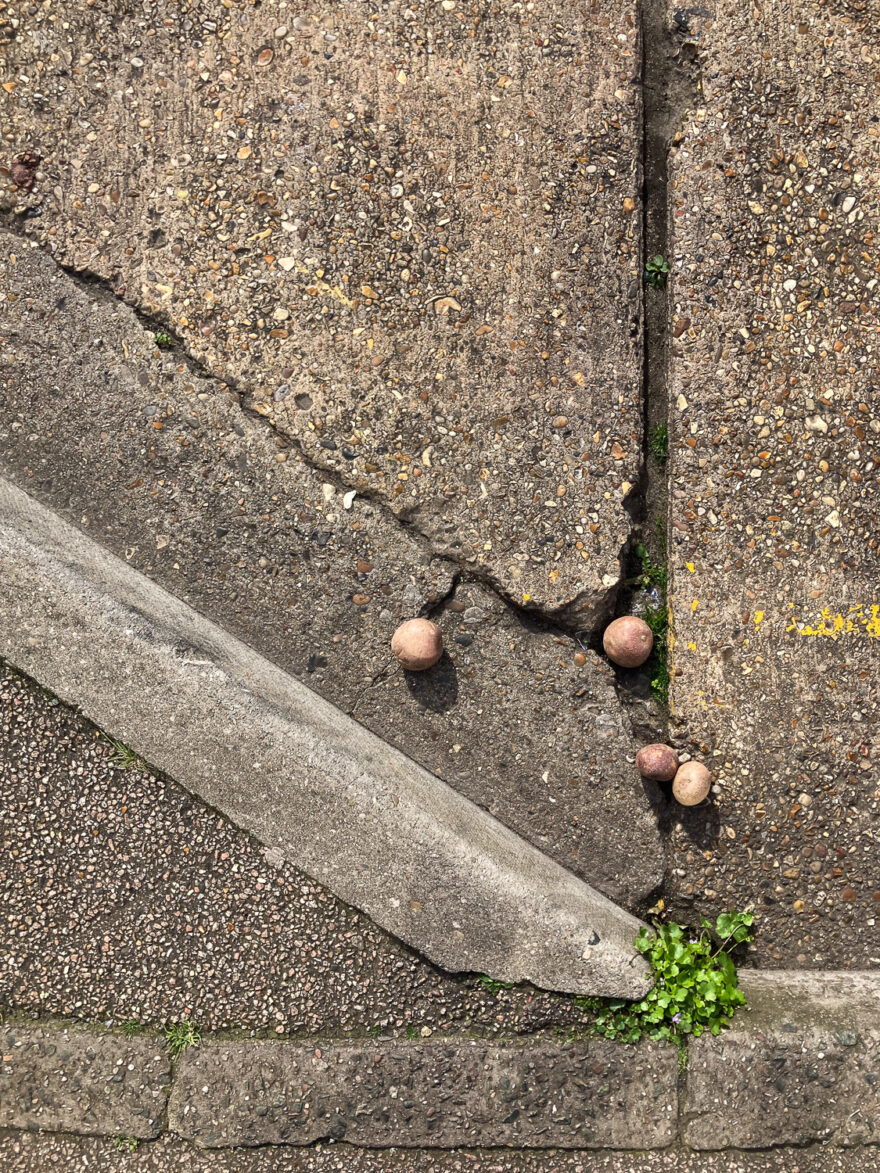 Photograph looking down at the ground, gutter at the side of the road running along the bottom and diagonally from the bottom right and up to the left. Four round potatoes are lying in the road, caught in the cracks.