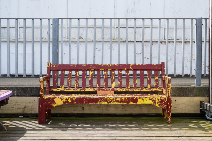 Photograph of a bench with weathered peeling dark red paint revealing yellow paint beneath, on a wooden floor with metal railings on a raised floor behind, with worn white walls behind that.