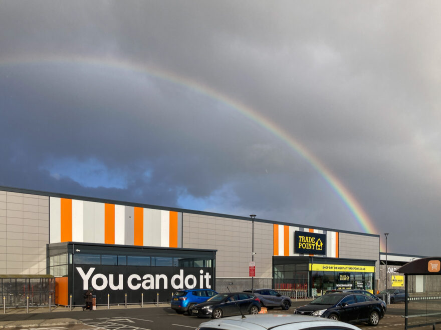 Photograph of a dark sky with a large, bright rainbow going from the top left and down to the right. Under the rainbow is a large shop with 'You can do it' written to the left and 'TRADE POINT' to the right. In front are cars parked in a carpark.