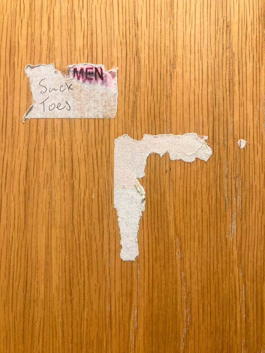 Photograph of a wooden surface with two partially removed white paper stickers on it, the one at top left has 'Suck Toes' hand written in black biro and 'MEN' in smudged red print to the right of that, slightly above.