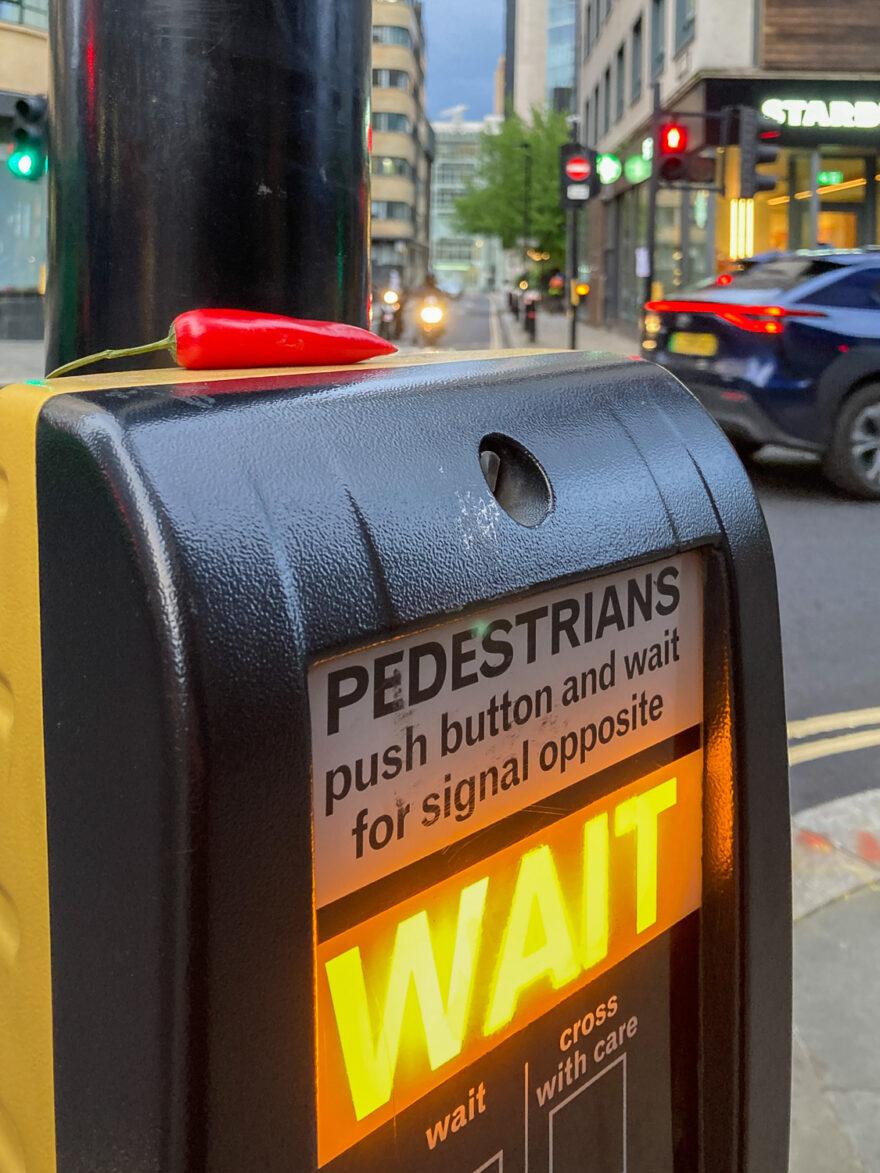 Photograph of a pedestrian crossing box with a red chilli pepper placed on the top. The text on the box says 'PEDESTRIANS, push button and wait for signal opposite' with 'WAIT' illuminated in yellow below and 'wait, cross with care' under that. In the background roads, shops and traffic can be seen out of focus.