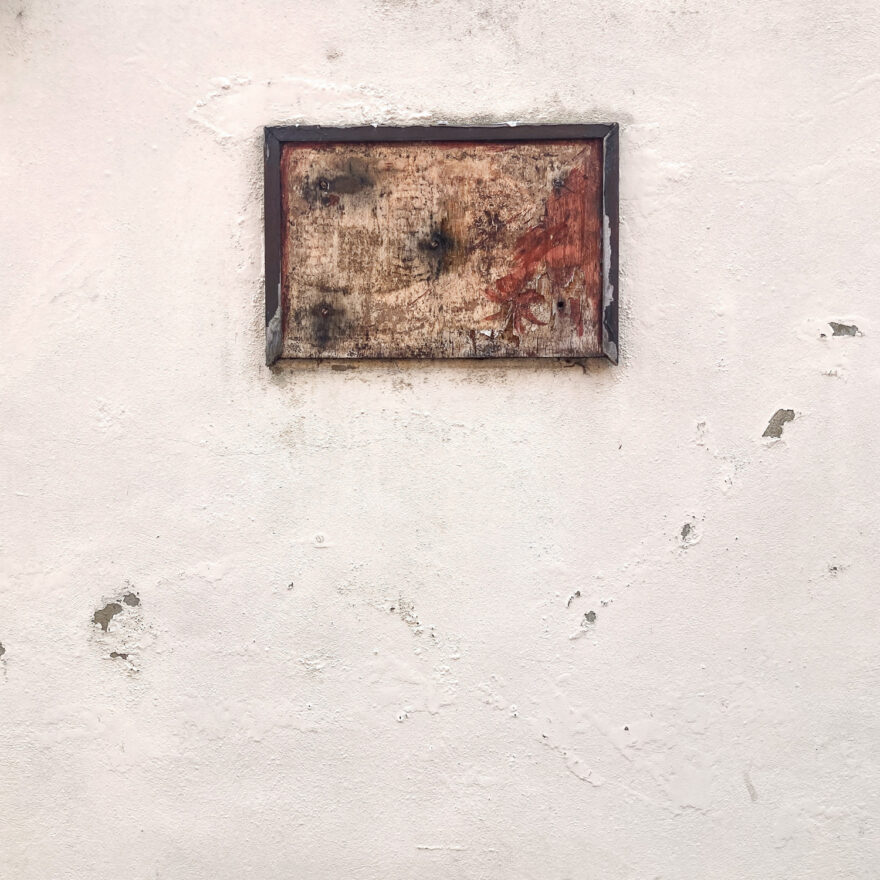 Photograph of weathered, broken wooden frame with nothing framed in it, in the top portion of the frame. On a peach, rough wall with bubbled and flaked plaster.