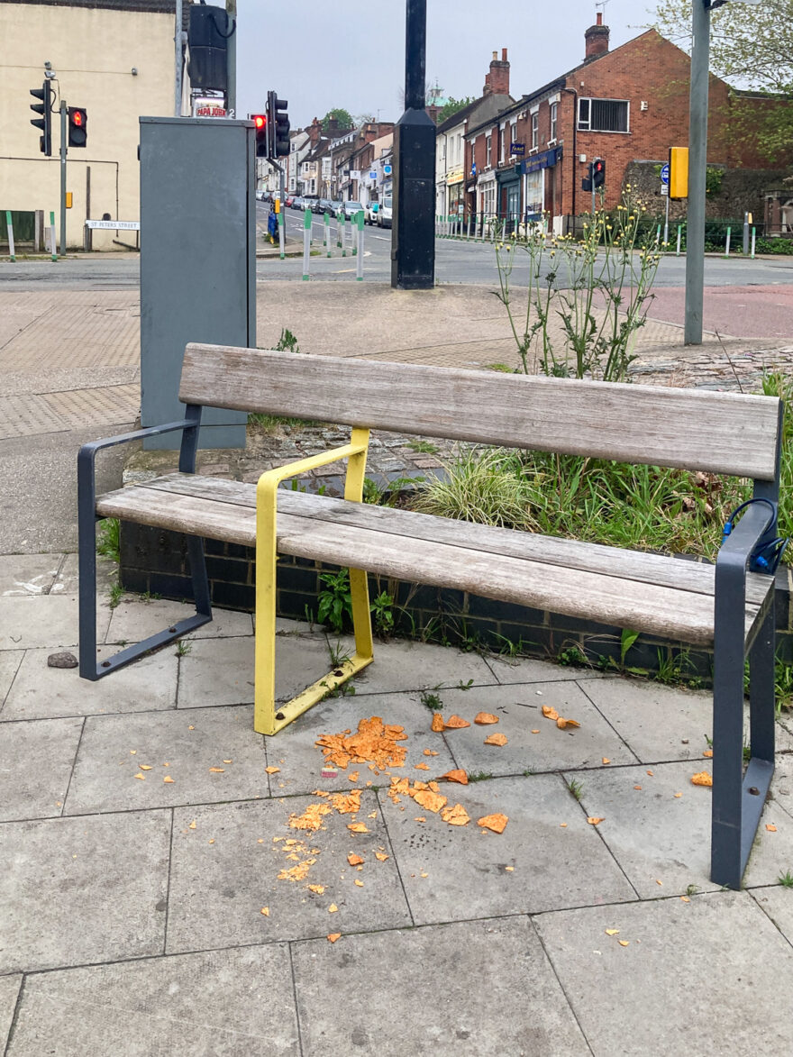 Photograph of a wooden and metal bench on a paved surface with road, buildings, crossing and planting area behind it. The bench has a yellow divider making one seat to the left and two seats to the right. Under the bench a load of tortilla chips have been dropped and broken on the floor.