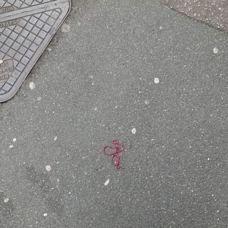 Photograph of a red strawberry lace sweet on a pavement, in a squiggly shape. There are remnants of chewing gum across the ground, a triangle of different tarmac-like material top right and part of a metal utility cover to the top left.