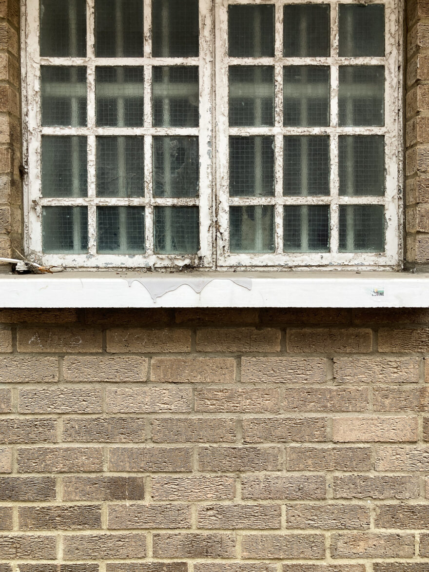 Photograph of dried dripping grey paint on a windowsill below a window with small reinforced panes with bars behind and brick wall below.