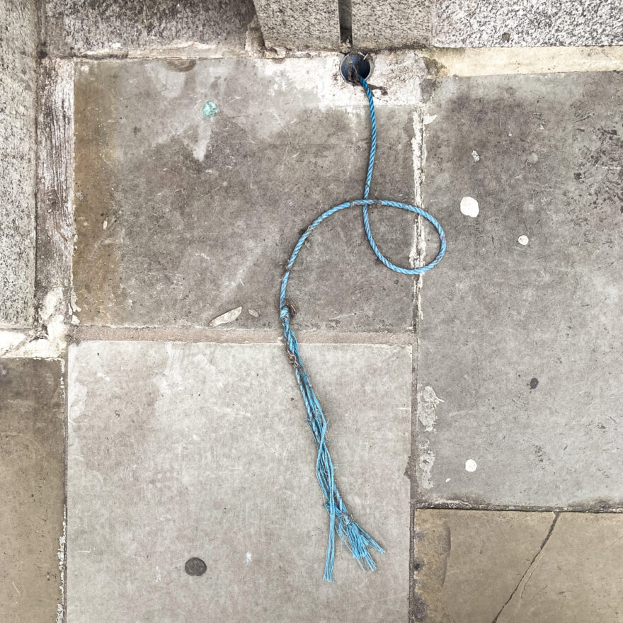 Photograph looking down at a paved pavement with a piece of blue rope lying on it in a loop, coming from a pipe in the ground at the top of the frame.