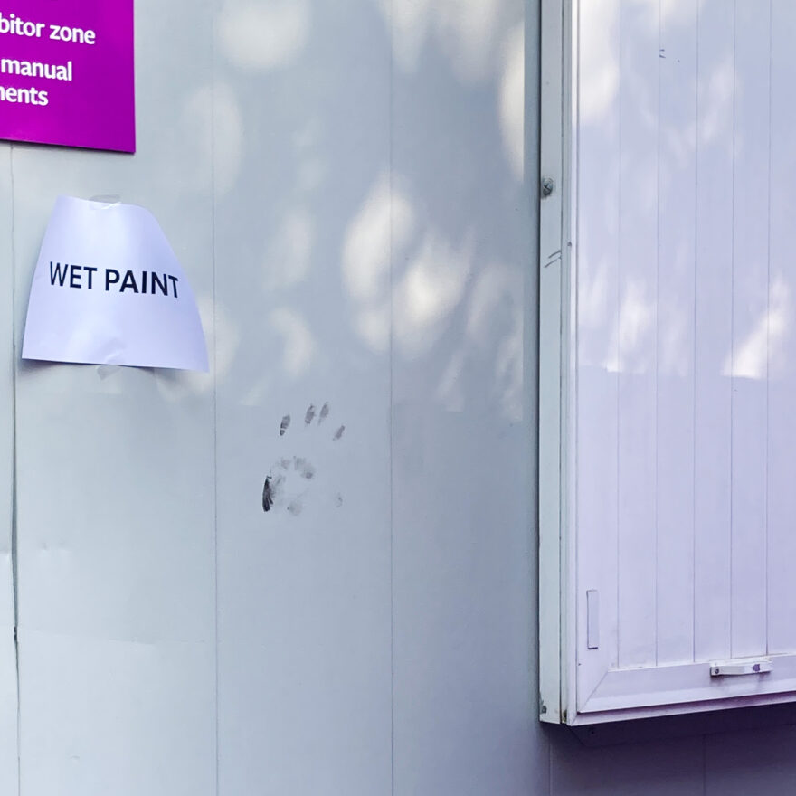 Photograph of a grey wall with a black handprint in the centre, dappled sunlight above, part of a purple sign to the top left, an A4 printed sheet taped below that saying 'WET PAINT'. Part of a white sign/window is visible to the right and a small amount of metal barrier can be seen at the bottom.