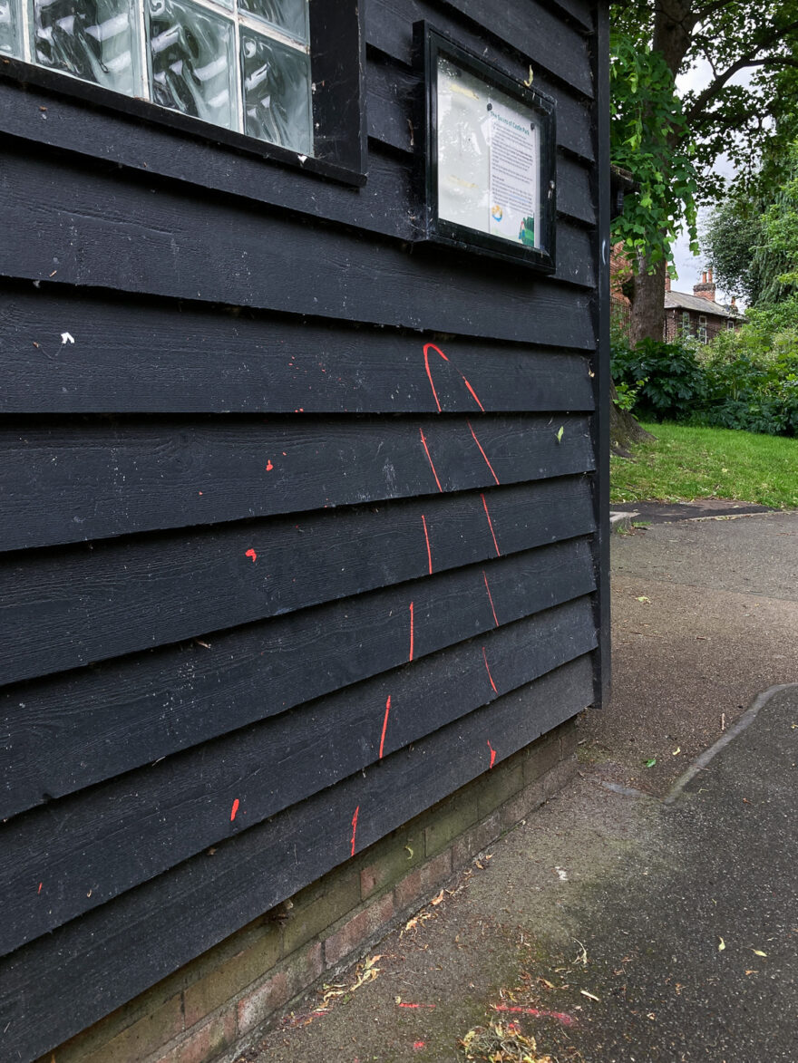 Photograph of a black wooden building with red paint squirted on it in a thin line up and curving back down. The line is broken across the planks of wood. There's a notice in a frame and part of a window visible at the top of the frame and to the right and behind is a concrete path, lawn, plants and the top of a house.