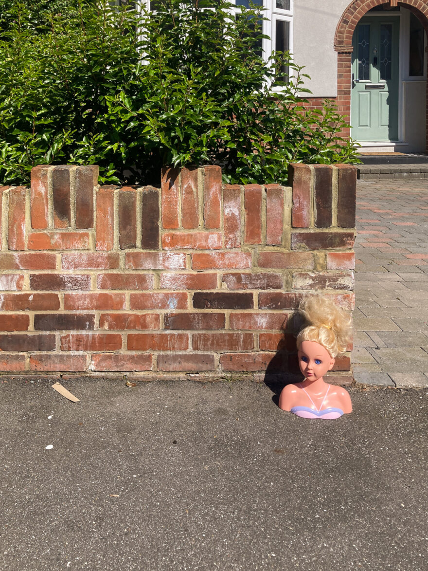 Photograph of a large doll's 'Girl's World' style head on a tarmac pavement next to a low brick wall with a green bush and front of a house with sage green front door behind.