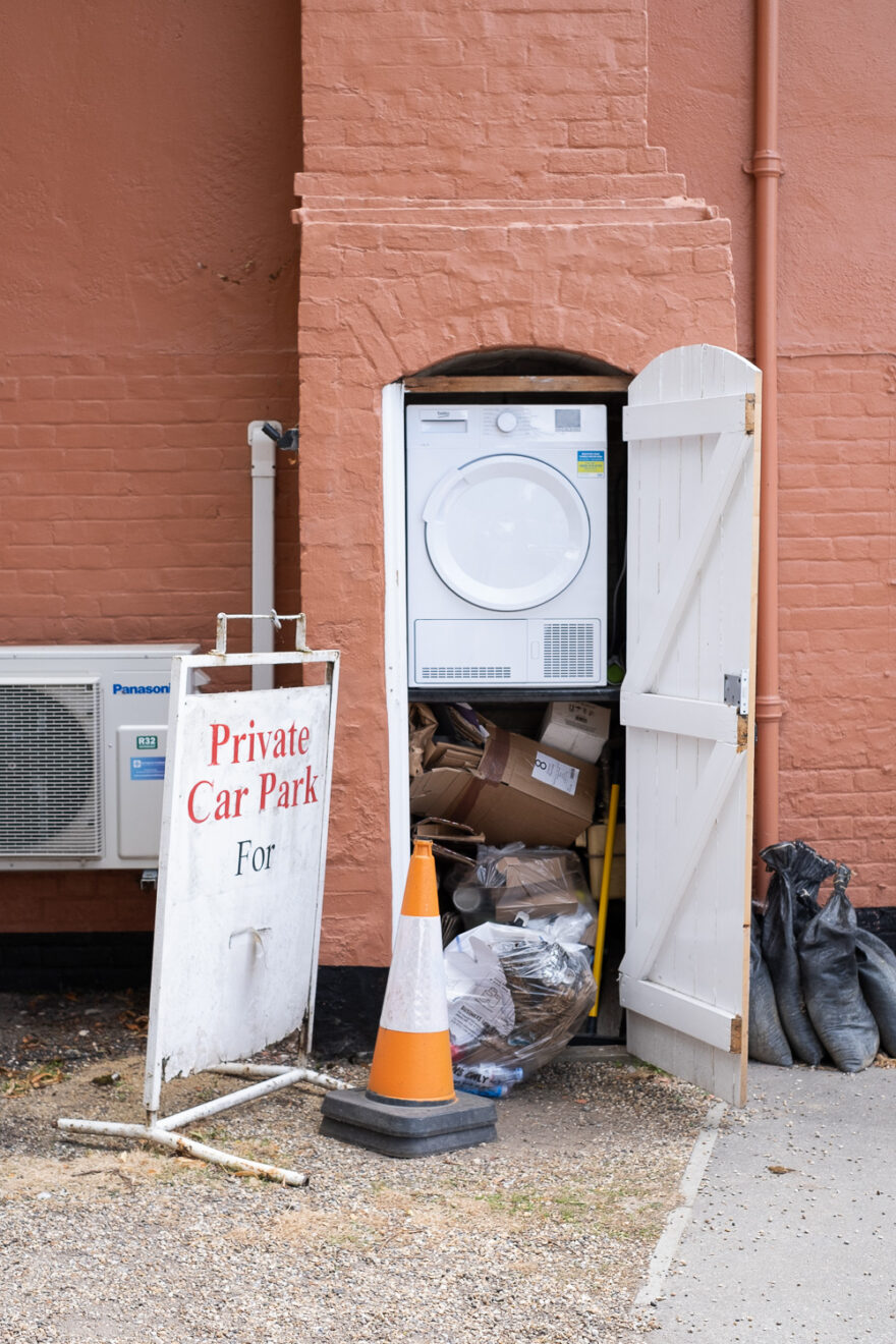 Photograph of a chimney breast with an open white wooden door showing a tumble drier inside at the top and empty cardboard boxes and rubbish below. The wall of the building is a salmon colour, there is a traffic cone, freestanding sign saying 'Private Car Park For' and an airconditioning unit to the left, gravel and concrete area in front and black sandbags lined up to the right.