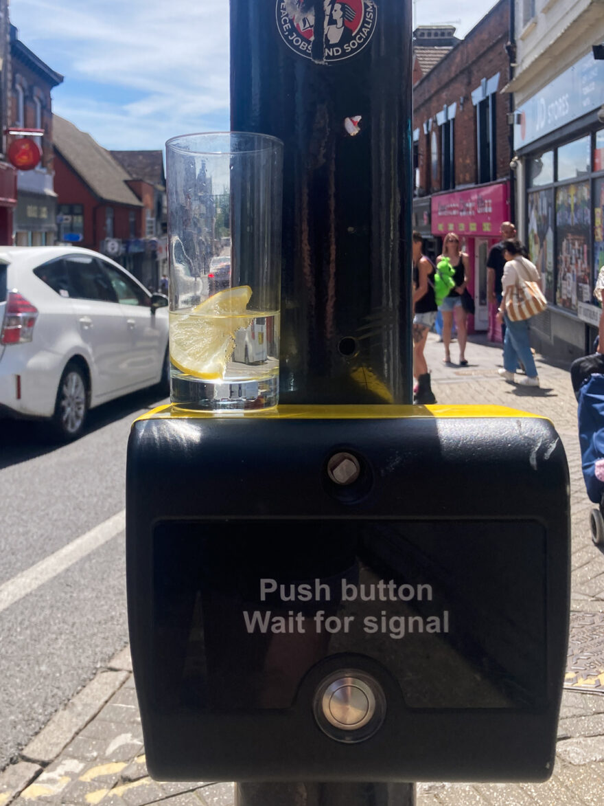 Photograph of a pole with a pedestrian crossing box that says 'Push button, Wait of signal' on it above a round metal button. On top of the box is highball glass with clear liquid and a slice of lemon in it. Behind is a street scene with a white car on the road to the left, people walking along the pavement in front of shops to the right.