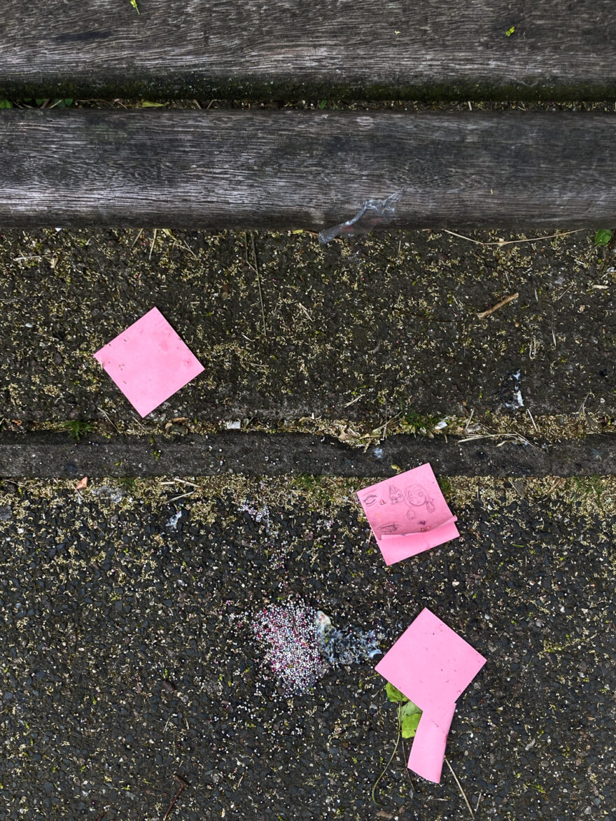 Photograph looking down a a tarmac pavement with pink post it notes in three sections, one in the centre with drawings on them. There is a melted icecream and sprinkles in the lower portion and wooden planks of a bench are in the top of the frame.