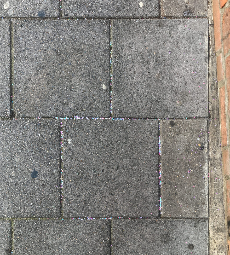 Photograph looking down at paving slabs with colourful glitter caught between the joins. To the right is a red brick wall.