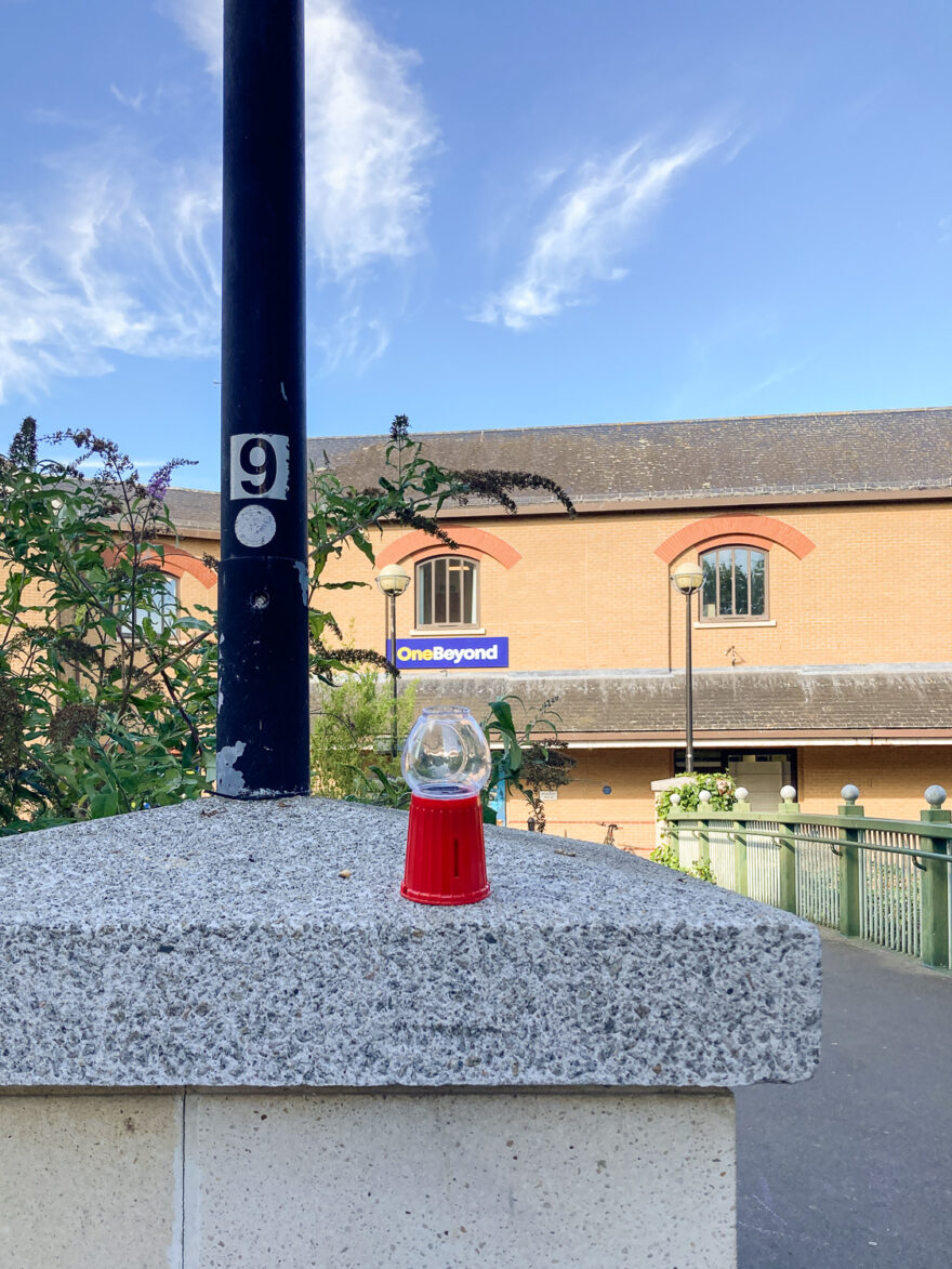 Chelmsford, UK, August 2024. Photograph looking down at paving slabs with colourful glitter caught between the joins. To the right is a red brick wall., UK, July 2024. Photograph of a small plastic empty gum ball machine on a concrete pillar, with a bridge across to a building behind, with a sign saying 'OneBeyond' on it, and deep blue sky with whispy clouds above.